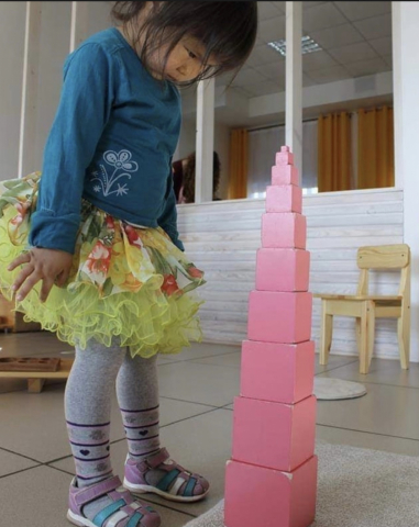 A young girl carrying Montessori Pink Tower cubes during a classroom activity A young girl carrying Montessori Pink Tower cubes during a classroom activity